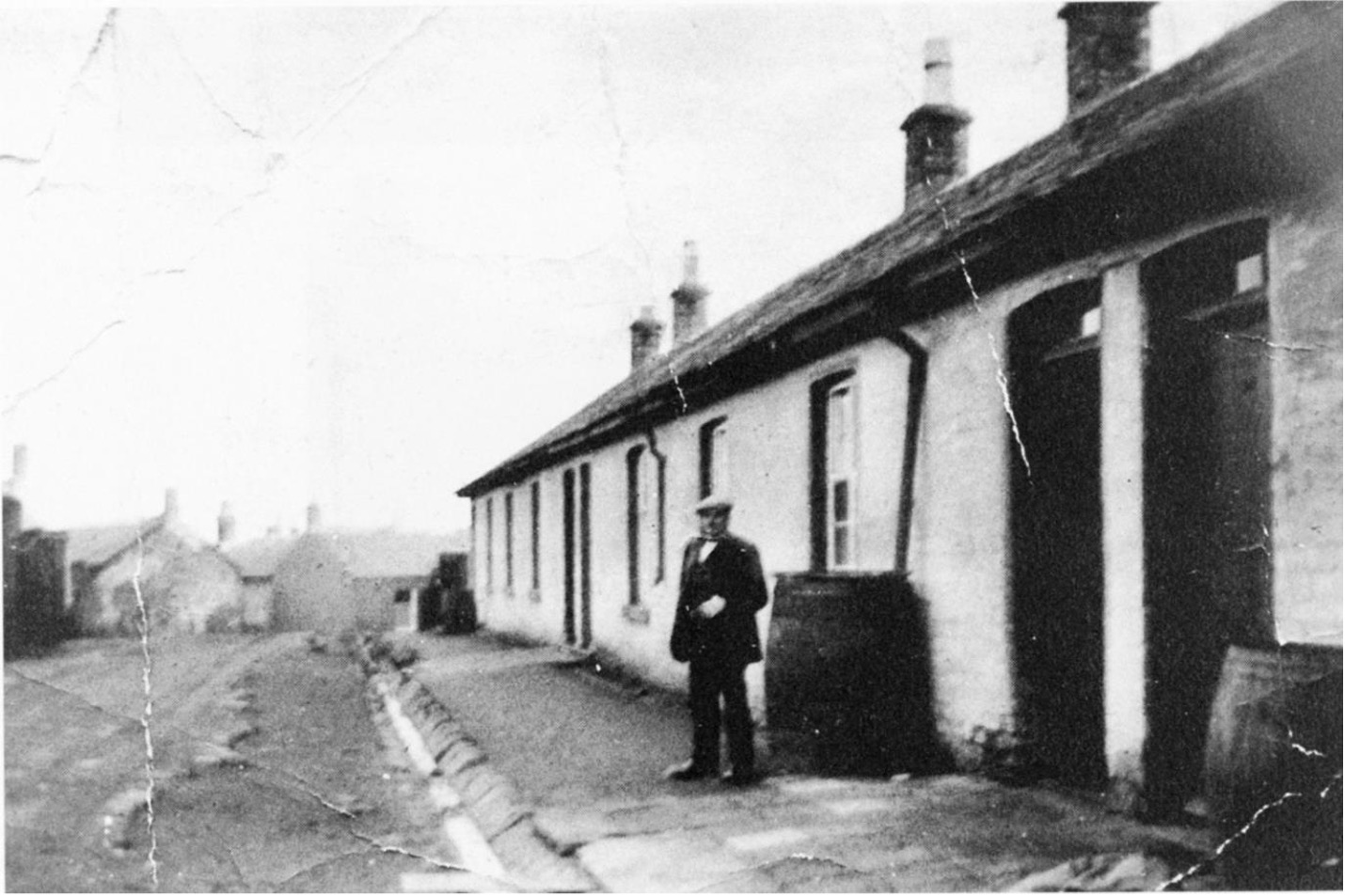 black and white photograph of a man wearing a dark suit and flat cap outside a single storey row of four houses which are painted white. Each house has a barrel under the downpipe. In the distances you can see more buildings.