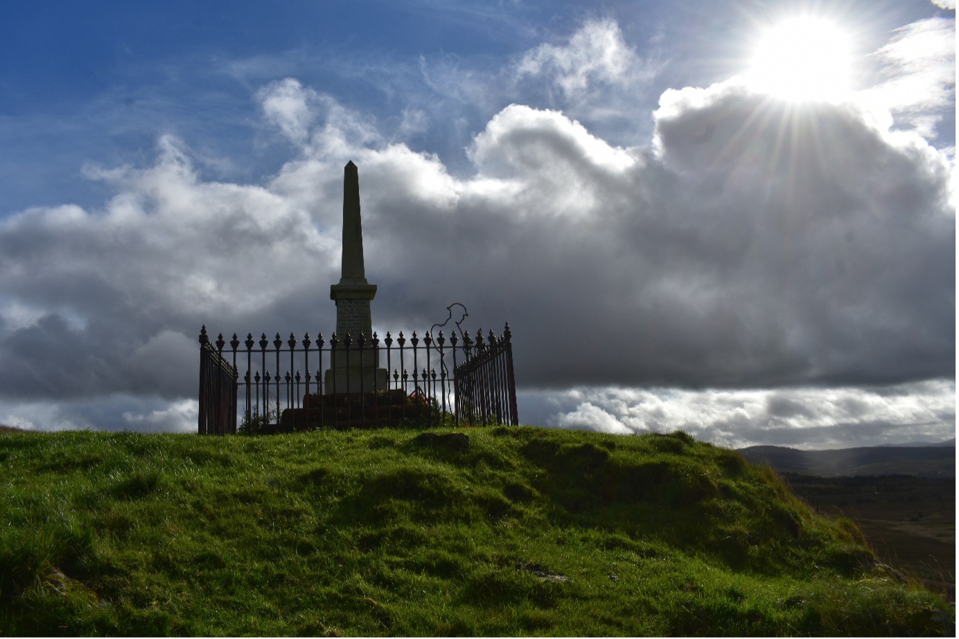 An obelisk shaped war memorial at Benwhat village Ayrshire. On a grass hill, with railing around the monument, silhouette of a Tommy solder next to he momument with a cloudy, blue sky and son in the top left hand corner