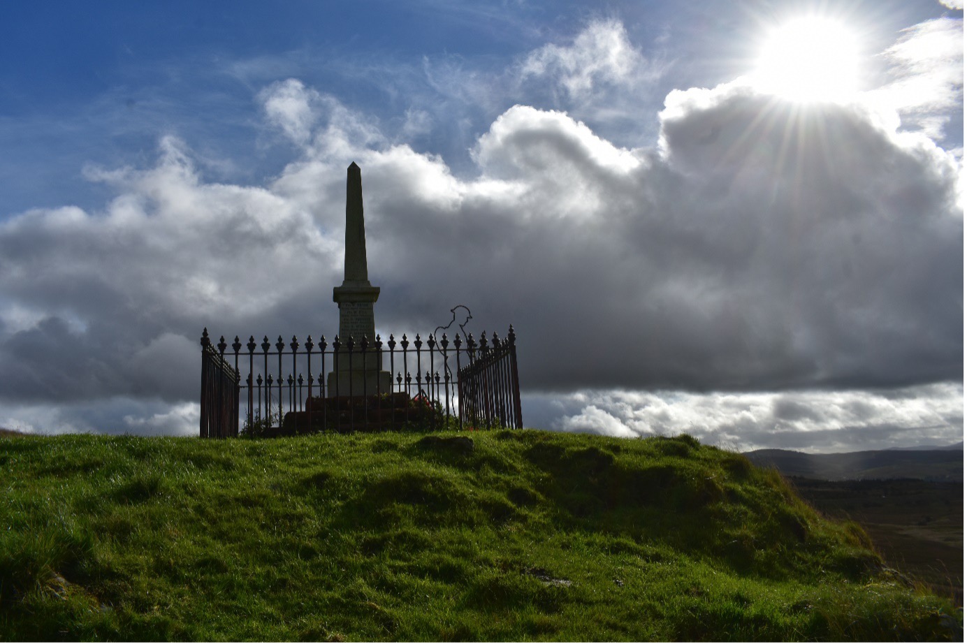 An obelisk shaped war memorial at Benwhat village Ayrshire.  On a grass hill, with railing around the monument, silhouette of a Tommy solder next to he momument with a cloudy, blue sky and son in the top left hand corner 