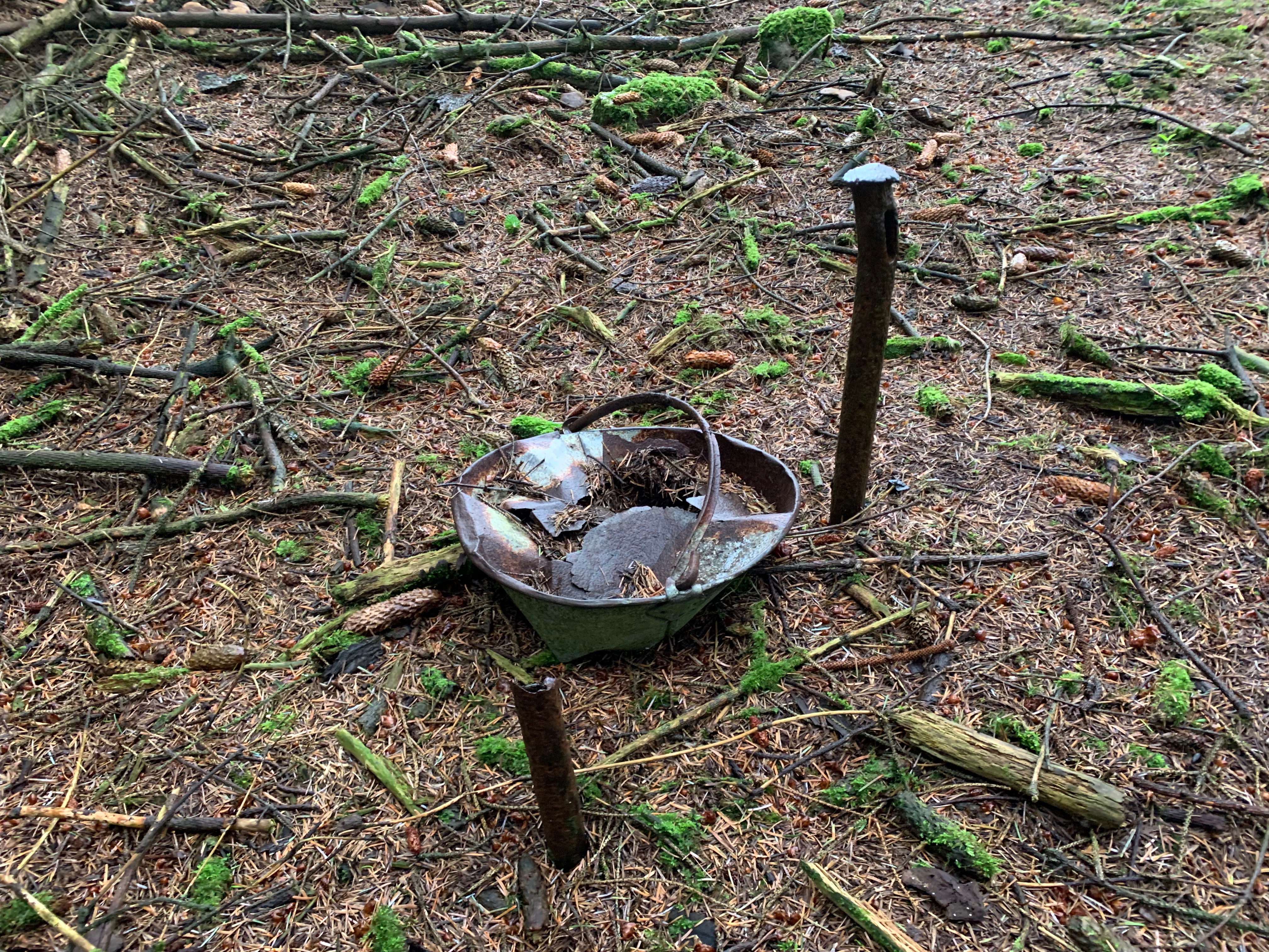 remains of a tin pail lid and handle next to some iron sticking out the ground of a forest floor