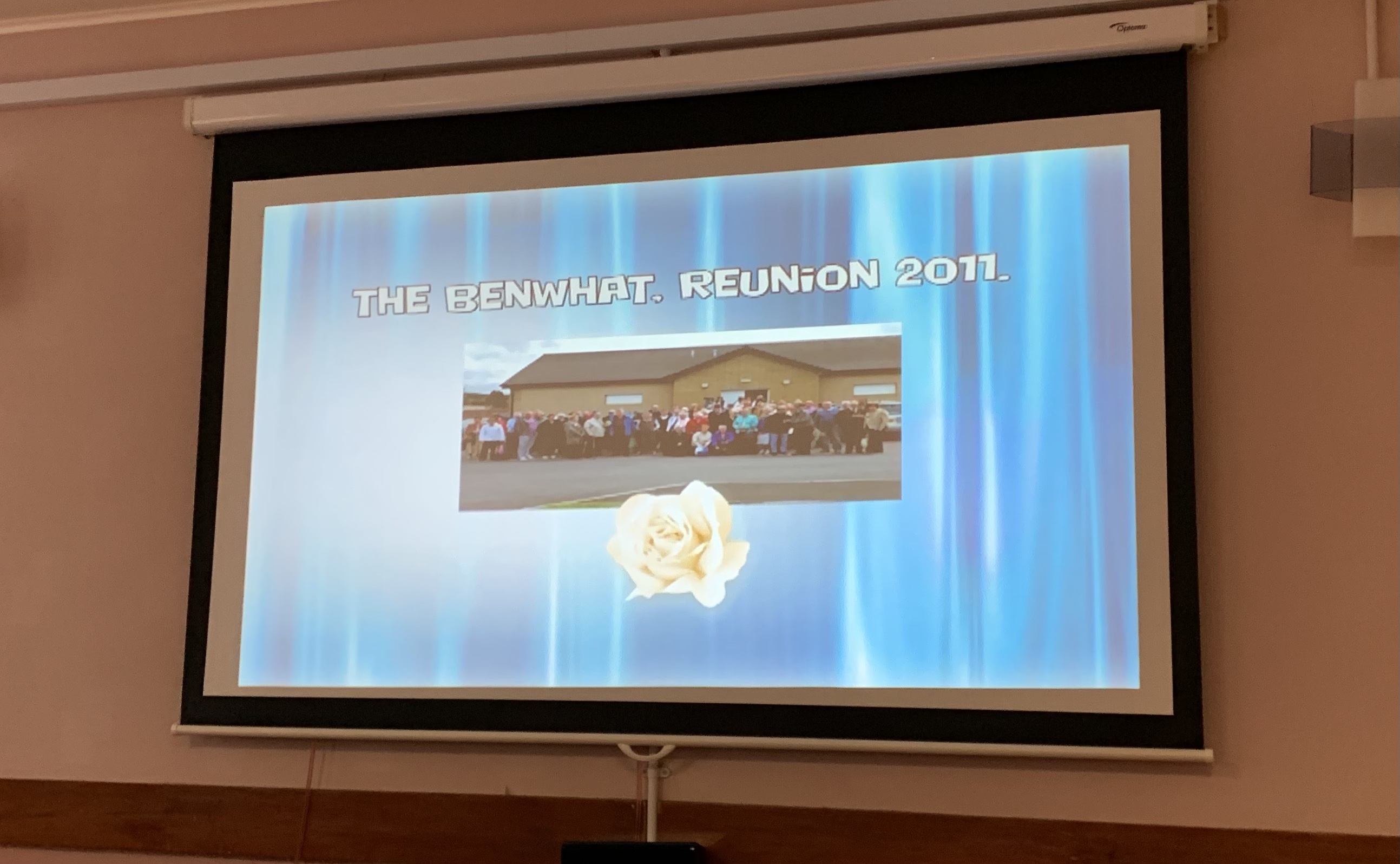 A screen on the wall showing a blue background with a photo in the centre. The words, The Benwhat Reunion, 2011, are along the top. 