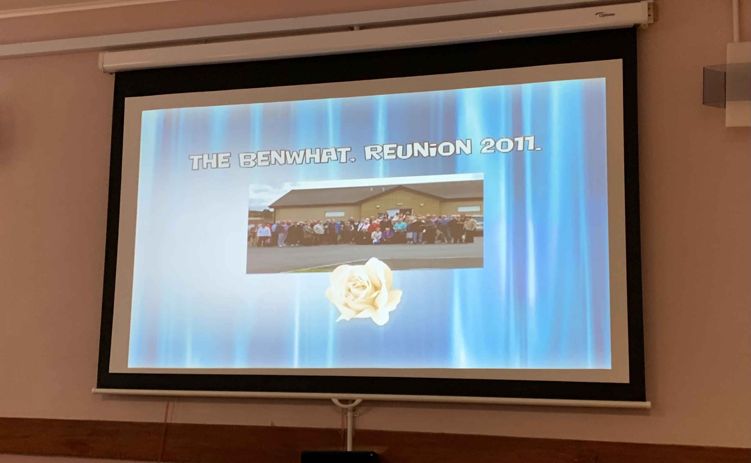 A screen on the wall showing a blue background with a photo in the centre. The words, The Benwhat Reunion, 2011, are along the top.
