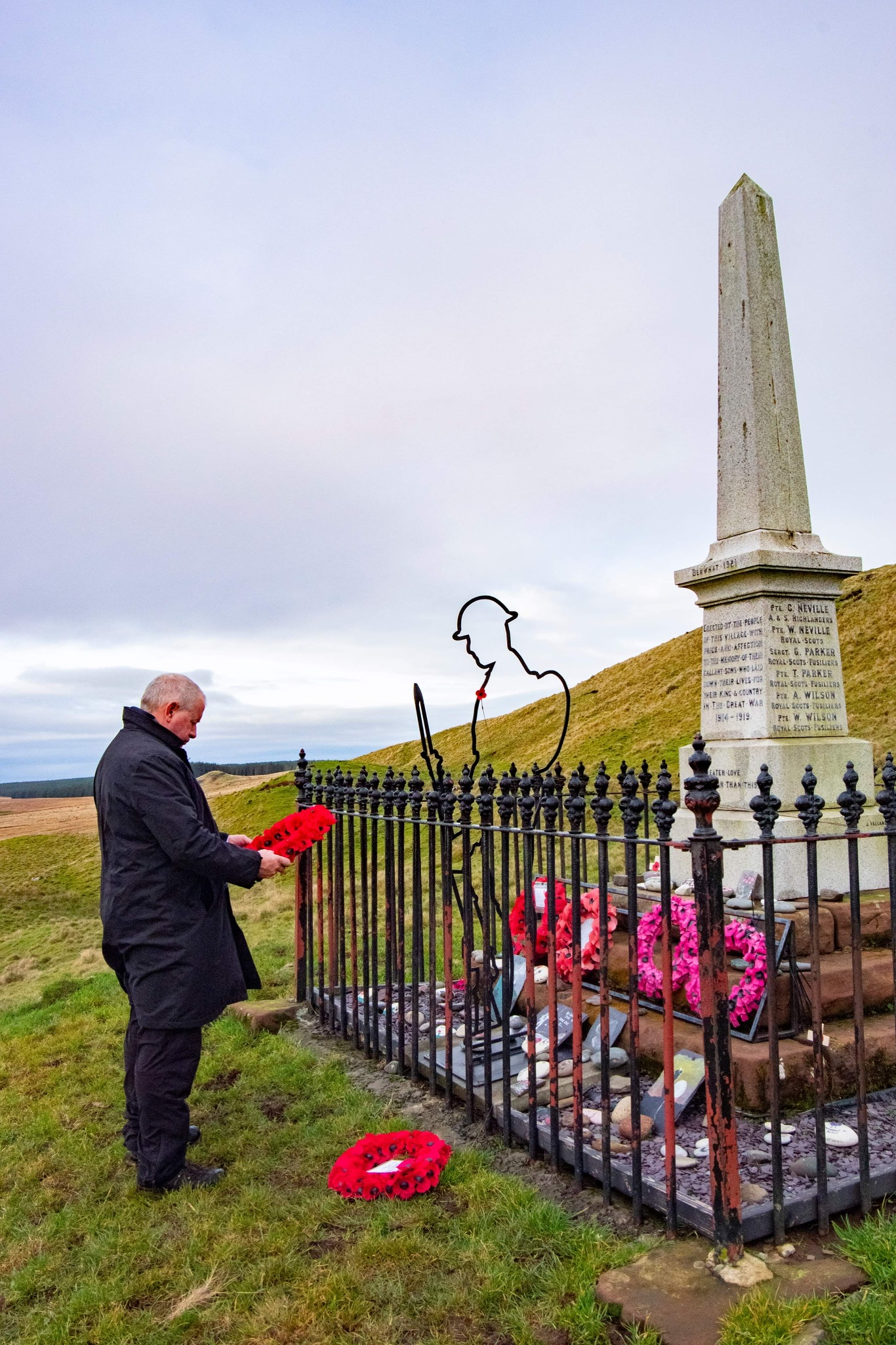 a man holding a poppy reef in front of the war memorial which is surrounded by black railings on a hillside