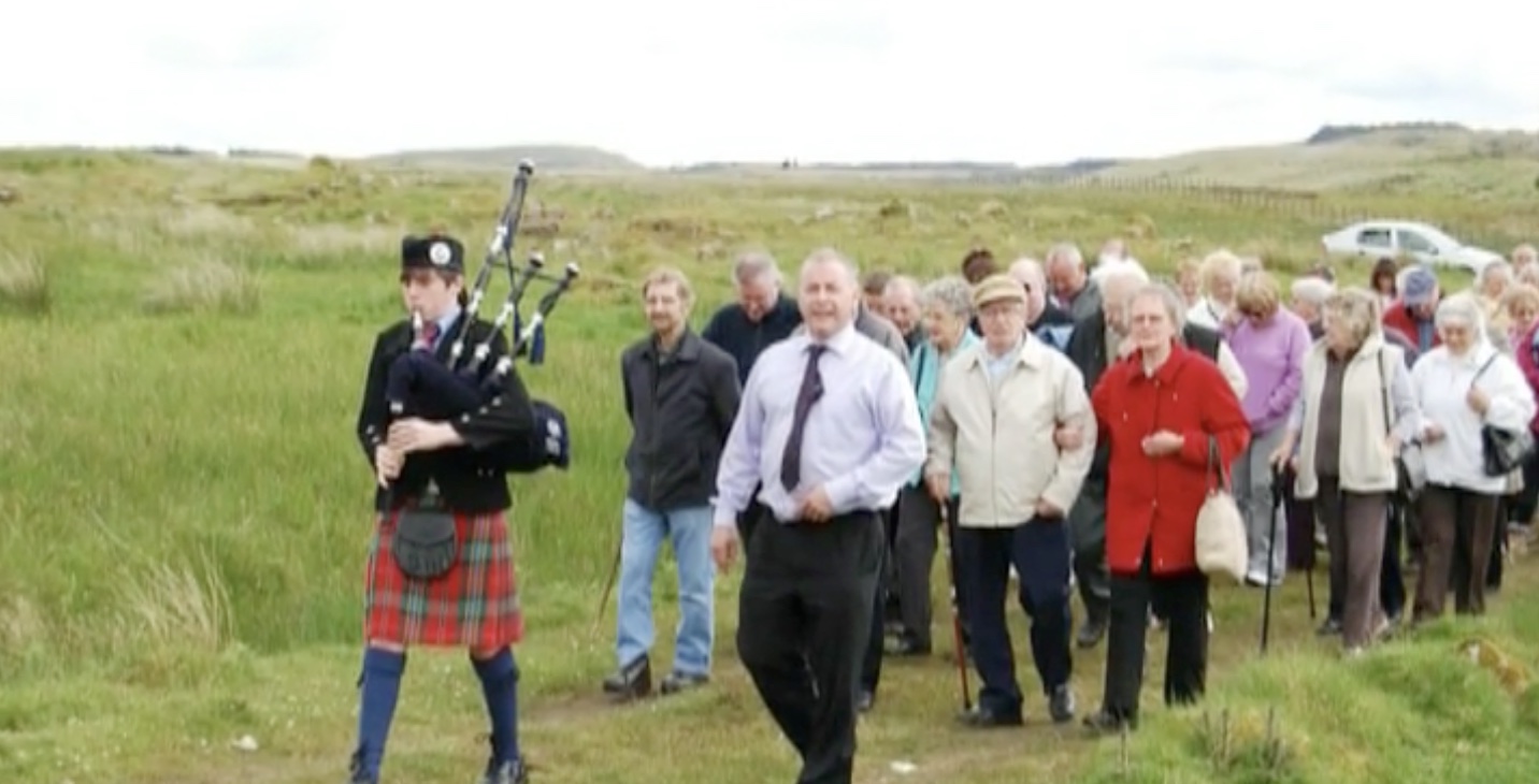 A group of people being led by a piper through the green hills of to the former mining village of Benwhat 