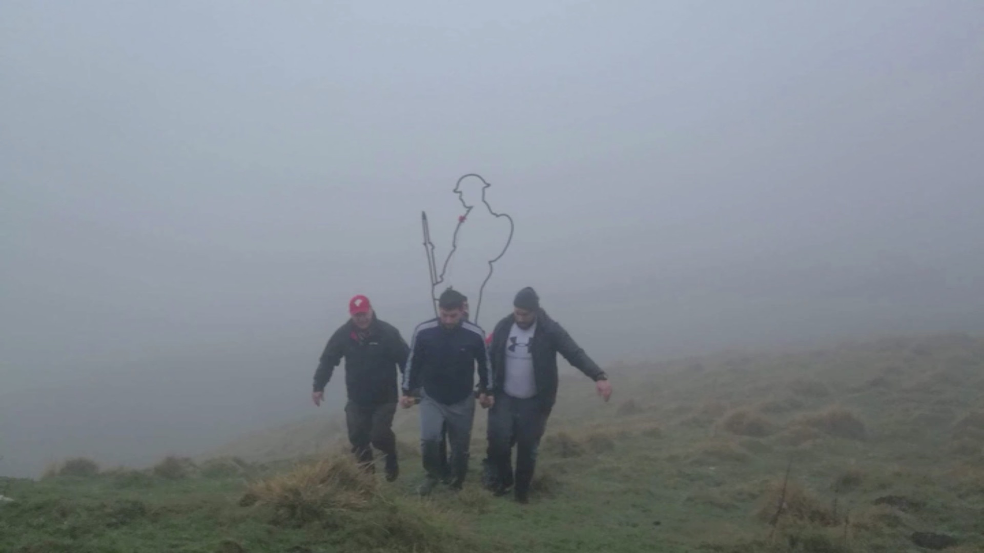 Three men caring a statue that is the outline of a World War Two Tommy carrying a rifle surrounded by mist on a hill