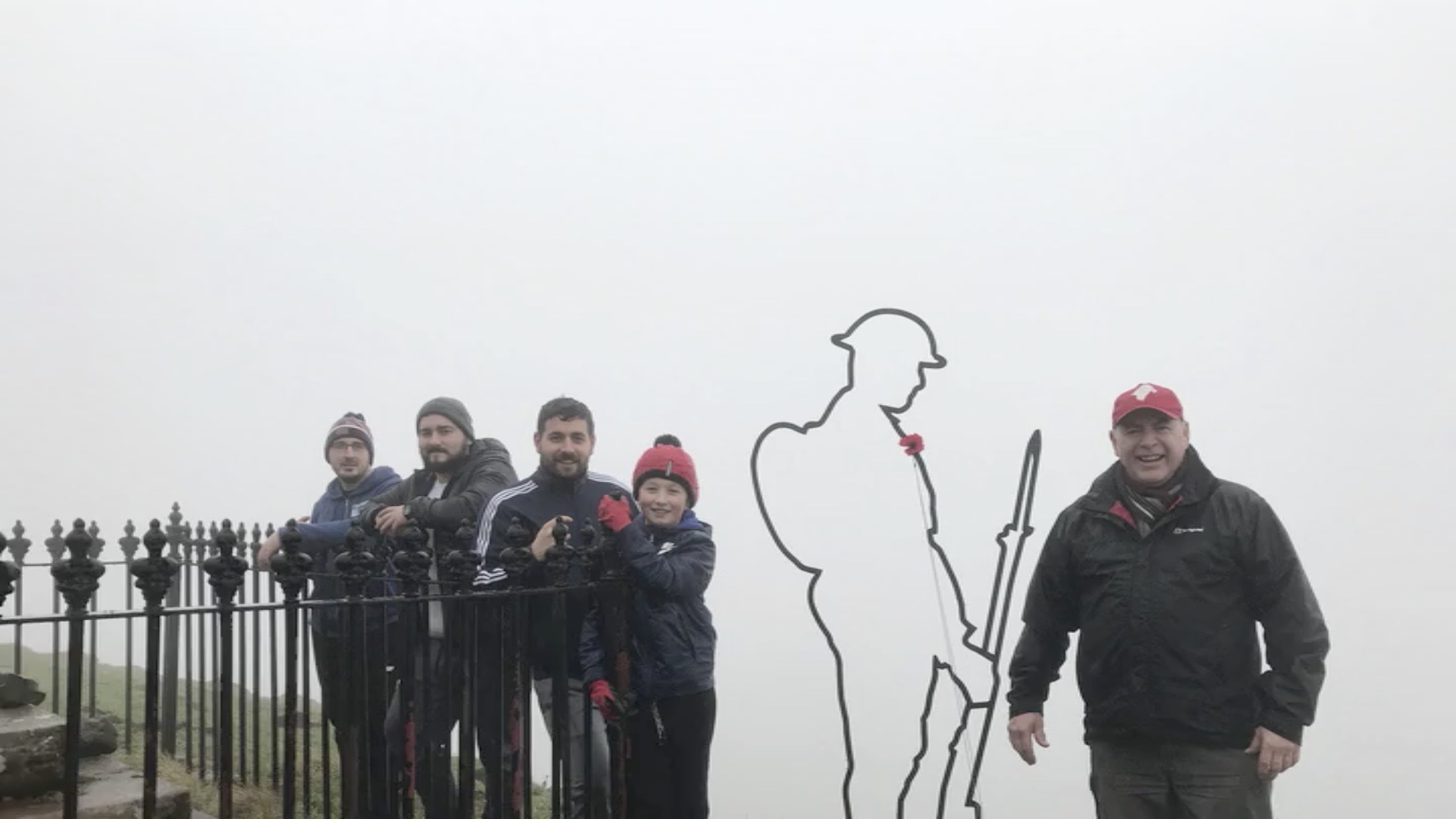 group of men with a young boy standing to the left of a statue which is an outline of a World War Two Tommy holding rifle. another man is standing on his own to the right of the statue