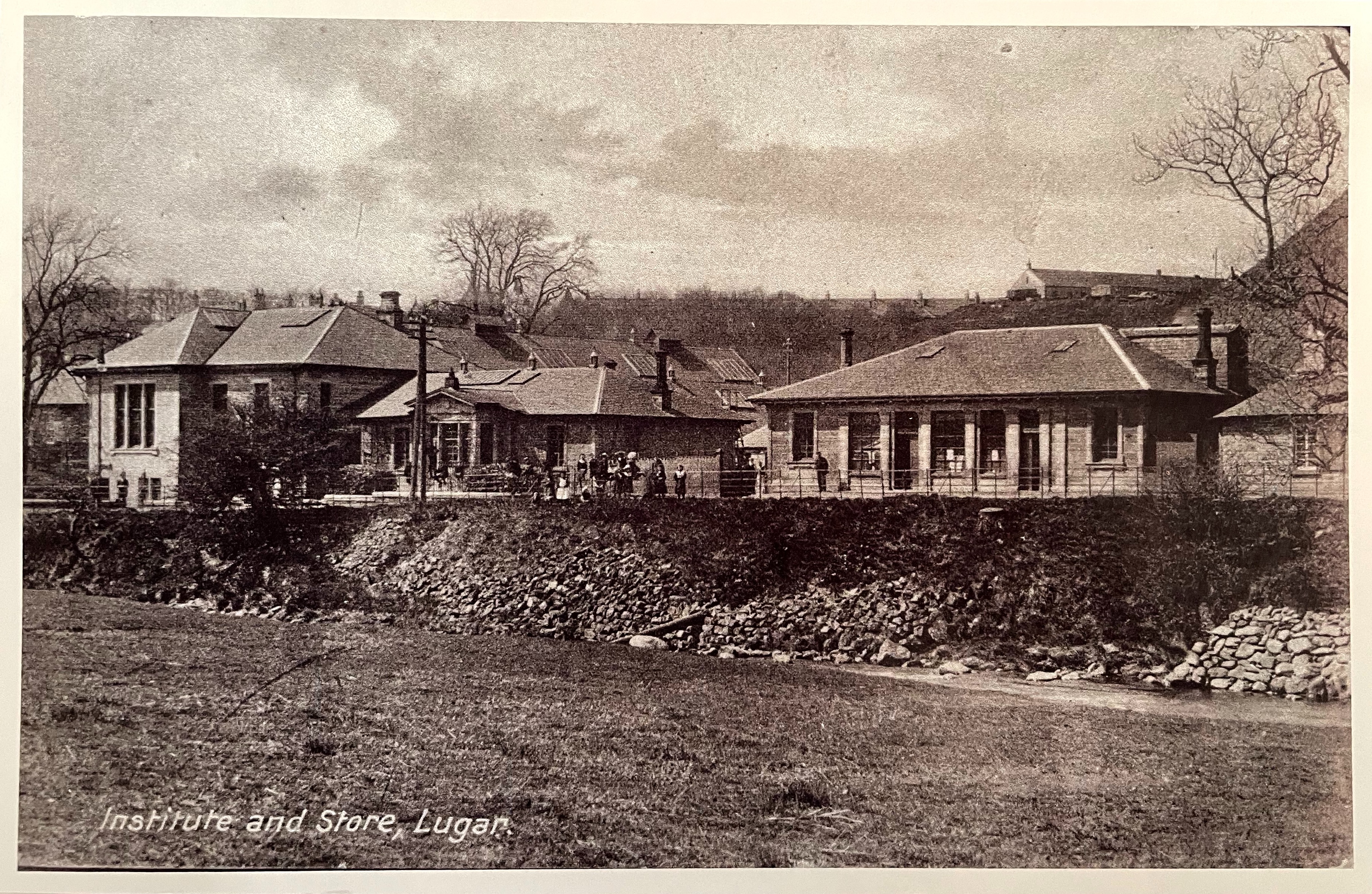 Postcard, black and white photo of a row of buildings with some grass in the foreground 