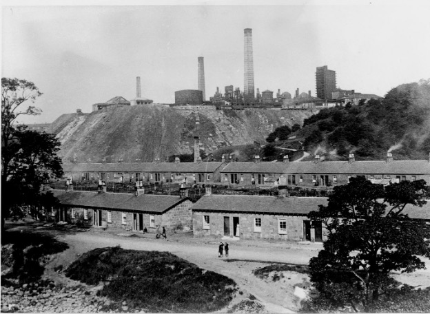 black ad white photography with two rows of houses with a ironworks towering over it in the background. The iron works has three chimney stacks.