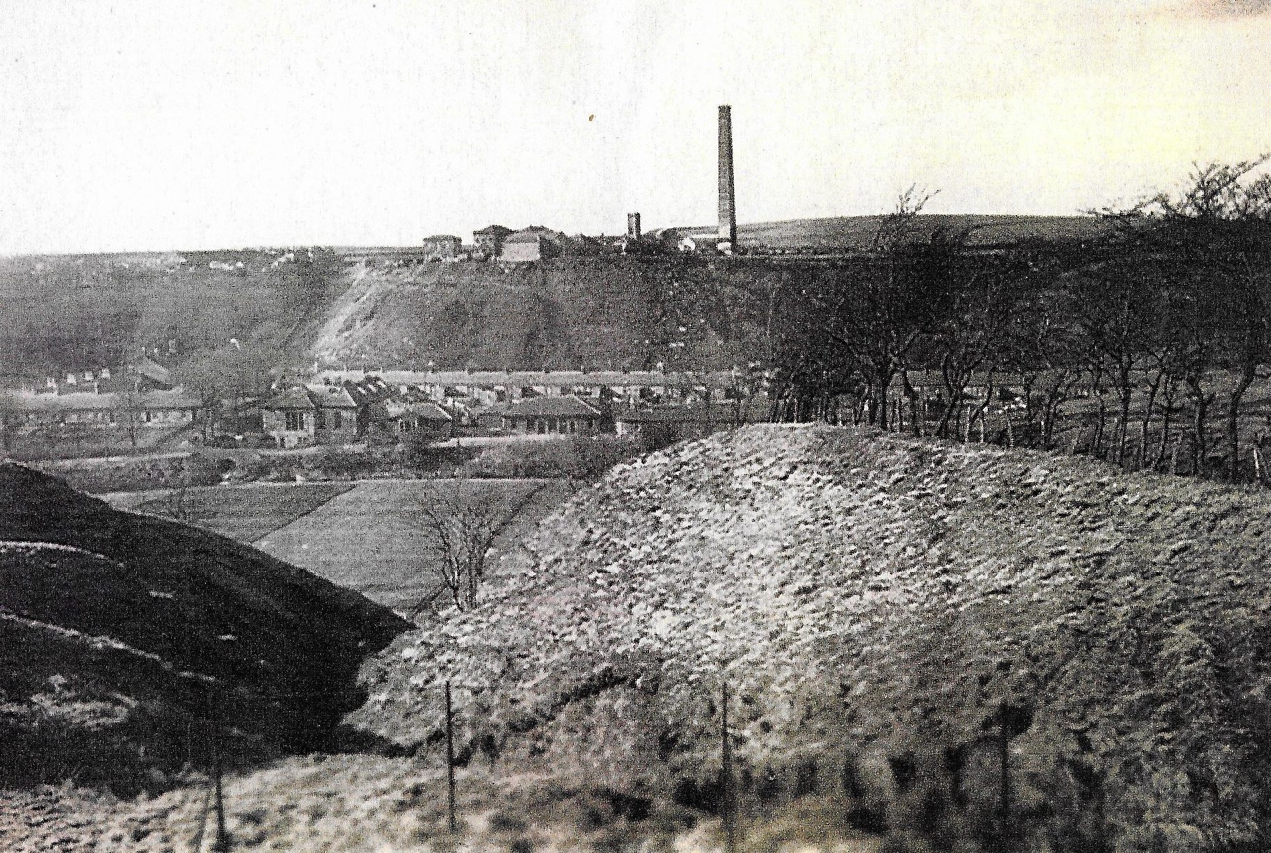 Black and white photograph of a mining village with the bing in the foreground, rows of houses in the middle and chimney stack in the background