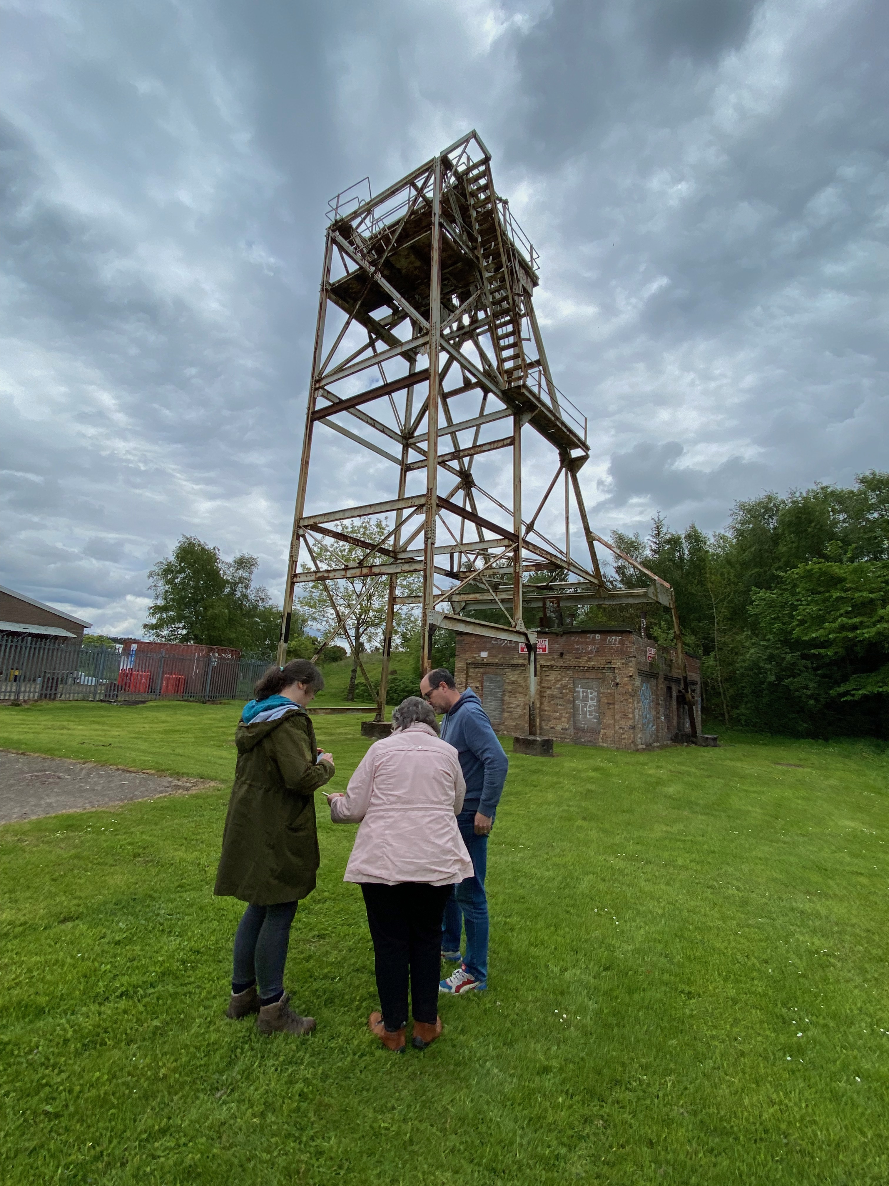 Three people, two female and one male standing looking at something in front of Highhouse mining wheel frame and steam house