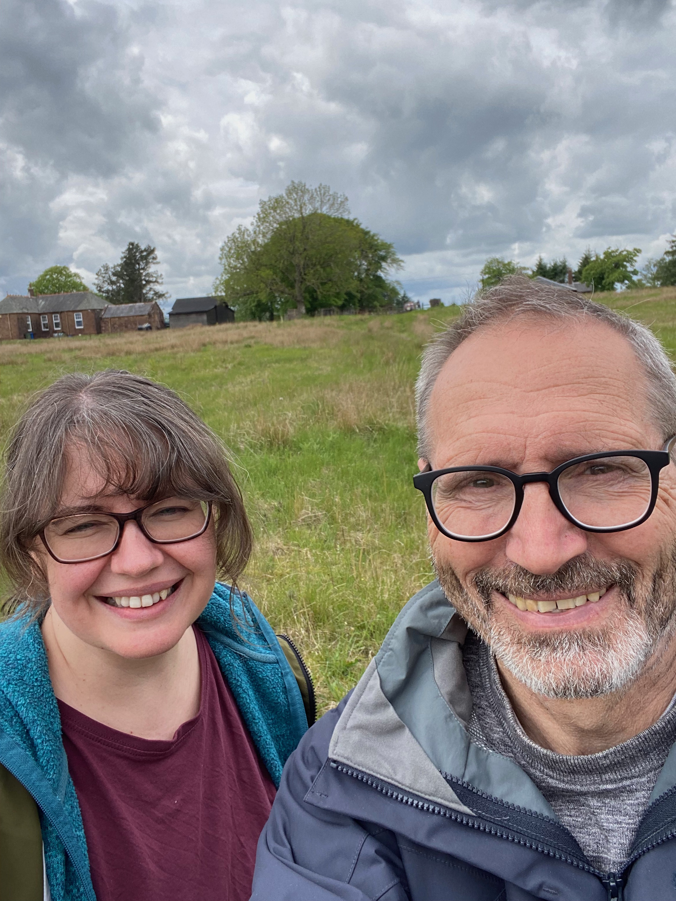 Two people wearing glasses, one male and one female, in a green field with a building in the background