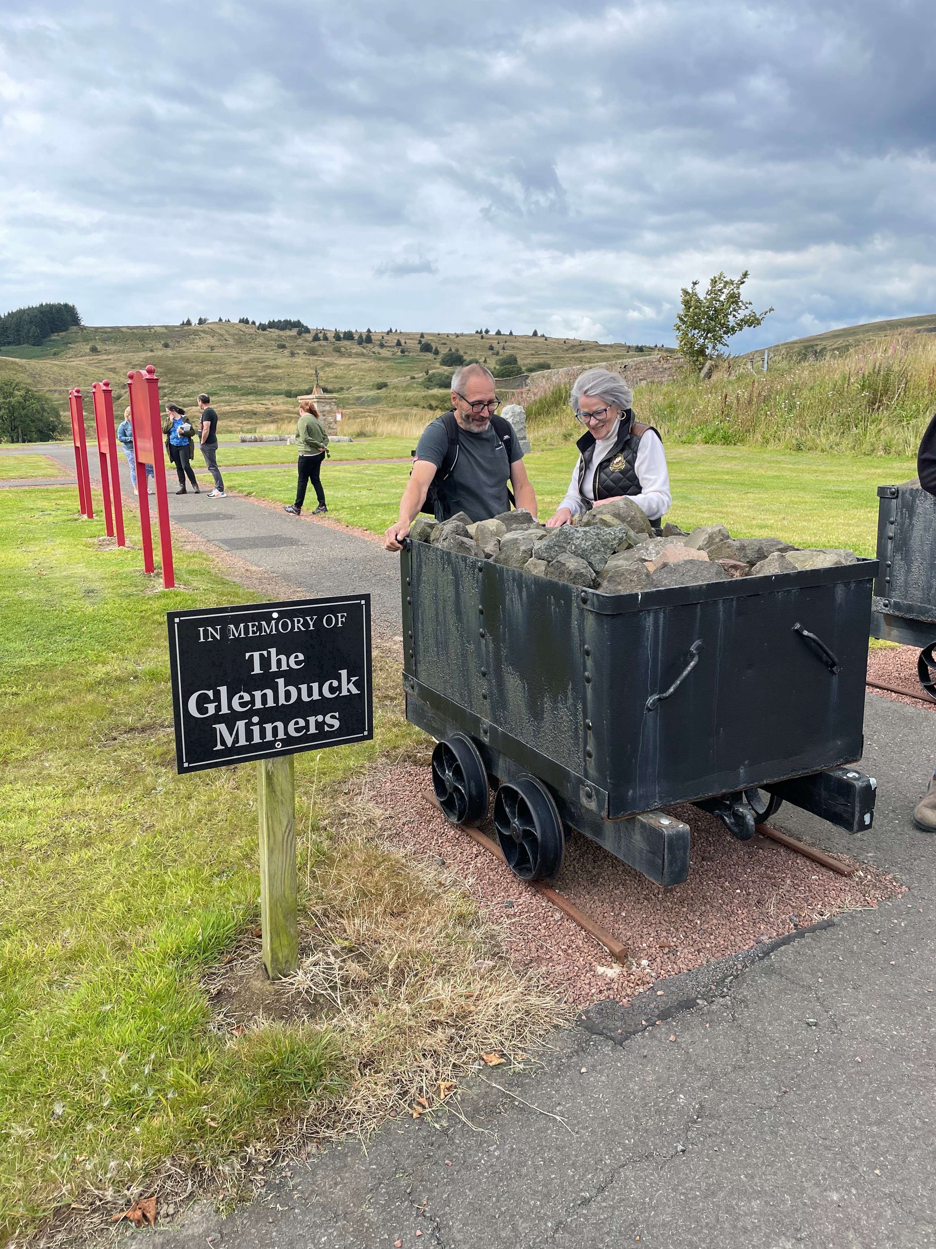 Two figures, one male and one female, looking a mining cart filled with stones next to a sign saying 'In Memory of the Glenbuck Miners'