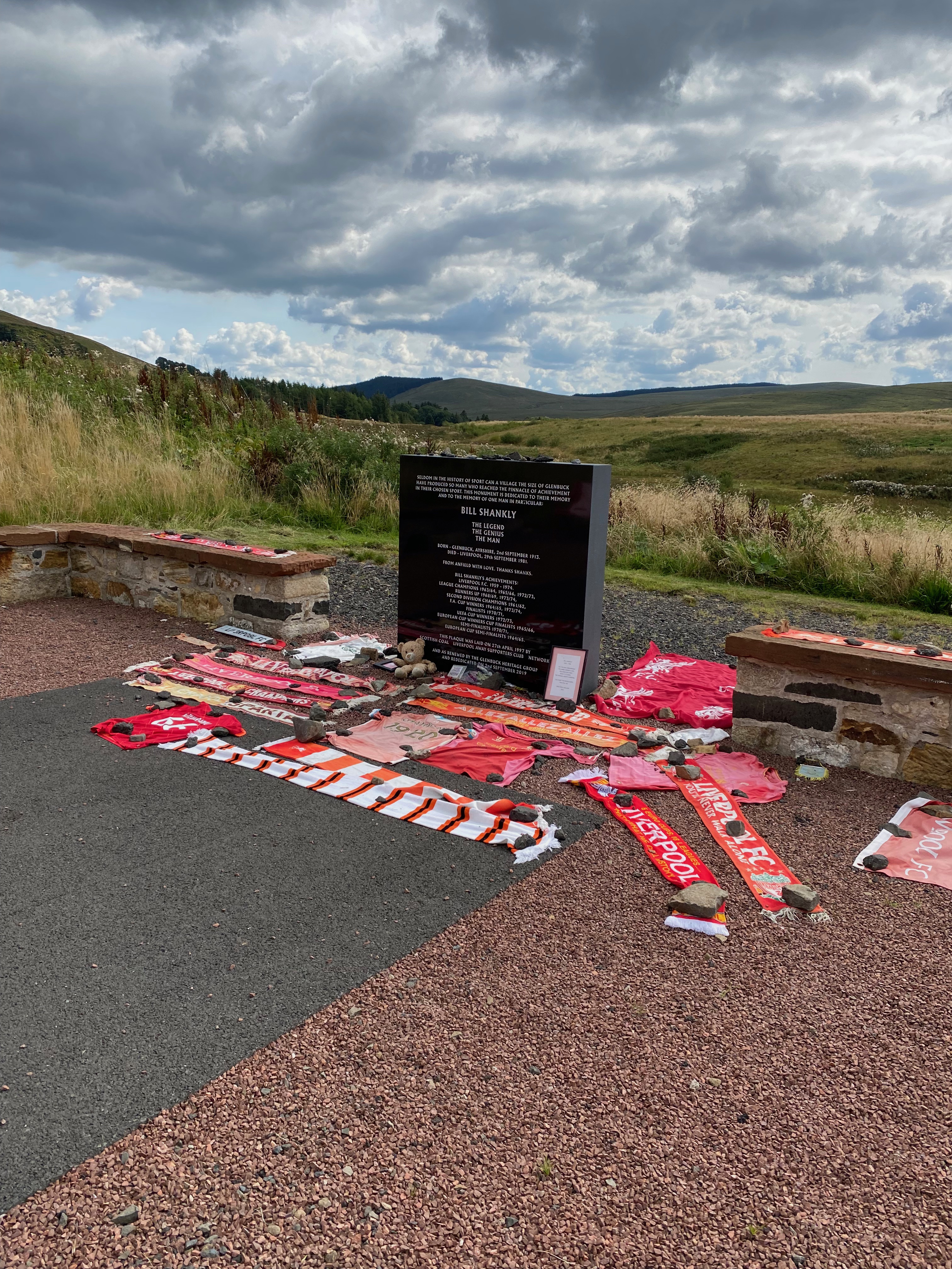 Black memorial stone surrounded by red and white scarves of Liverpool football club. Hills and a cloudy sky in the background