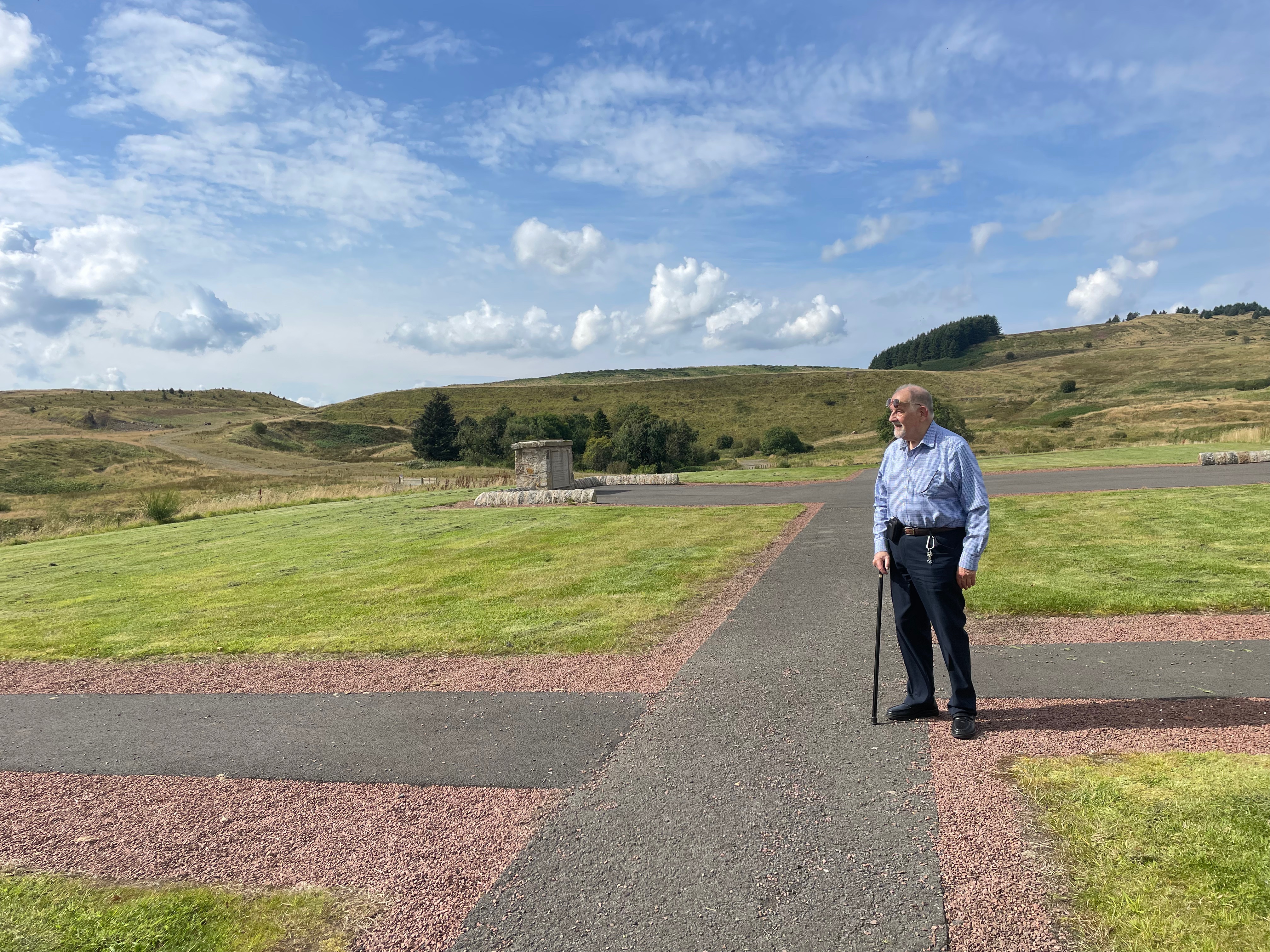 Man with a walking stick standing on some pathways surrounded by green grass and hills. 