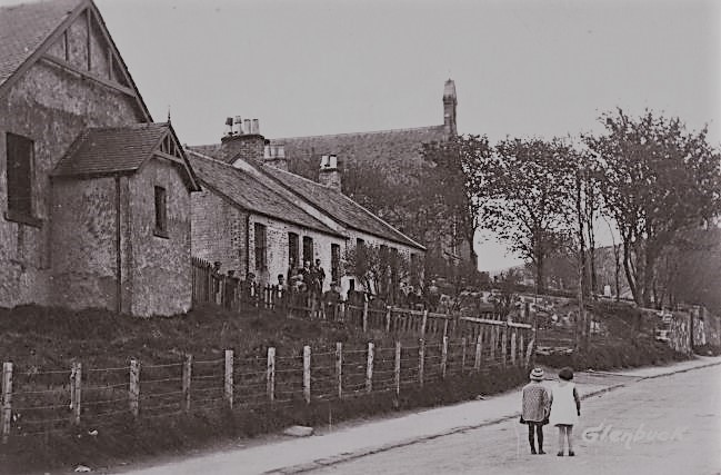 black and white photo of row of houses with a church in the background. Two children are in the street and and number of people on the doorstep of the first house 