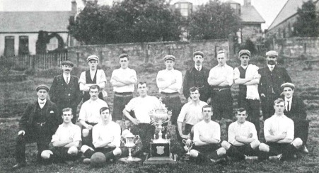 Photograph in black and white of a football team from the 1930s with two trophies
